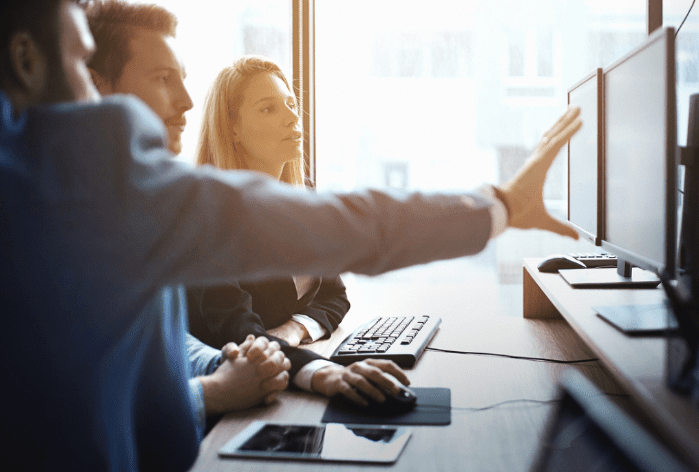 Two people focus on a computer monitor in a bright office, while a third person explains something, pointing at the screen. A keyboard and tablet are on the desk. Light from a window illuminates the scene.