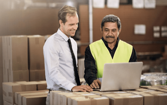 Two men stand in a warehouse in front of a laptop on a cardboard box. One wears a white shirt and tie, and the other wears a high-visibility vest. They both smile and focus on the laptop screen. Stacked boxes surround them.
