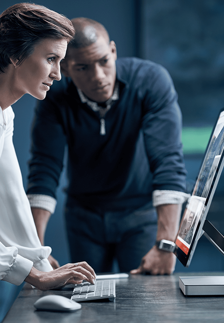 Two focused individuals are collaborating at a computer desk, examining a large monitor. One person is seated and typing, while the other is leaning forward. The setting appears professional and modern.