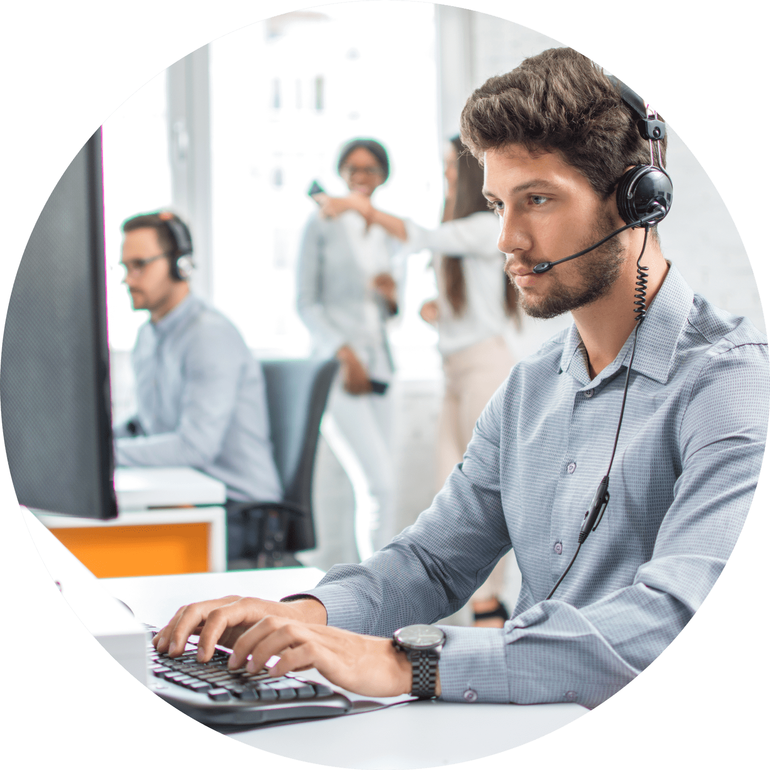 A man wearing headphones and a gray shirt is typing on a keyboard at a desk with a computer screen. Two colleagues are visible in the blurred background, speaking. The setting appears to be a modern office.