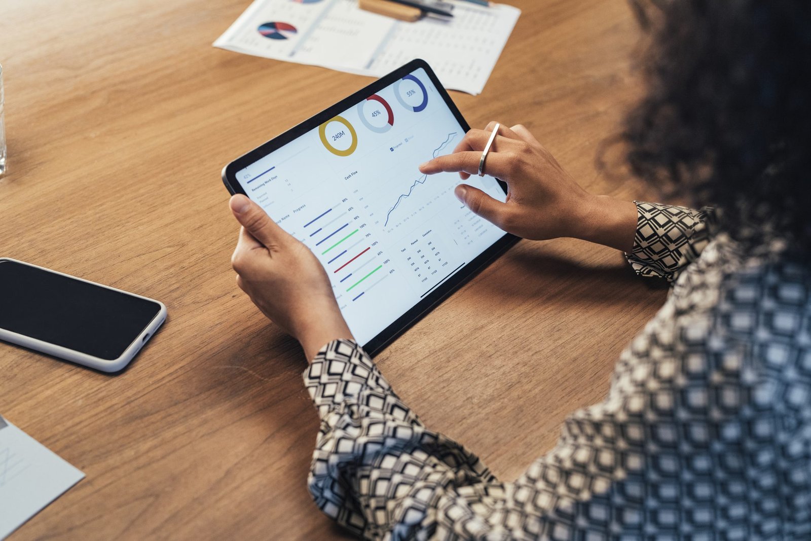 A person in a patterned shirt uses a stylus on a tablet displaying charts and graphs. The tablet is on a wooden table with documents and a smartphone nearby.