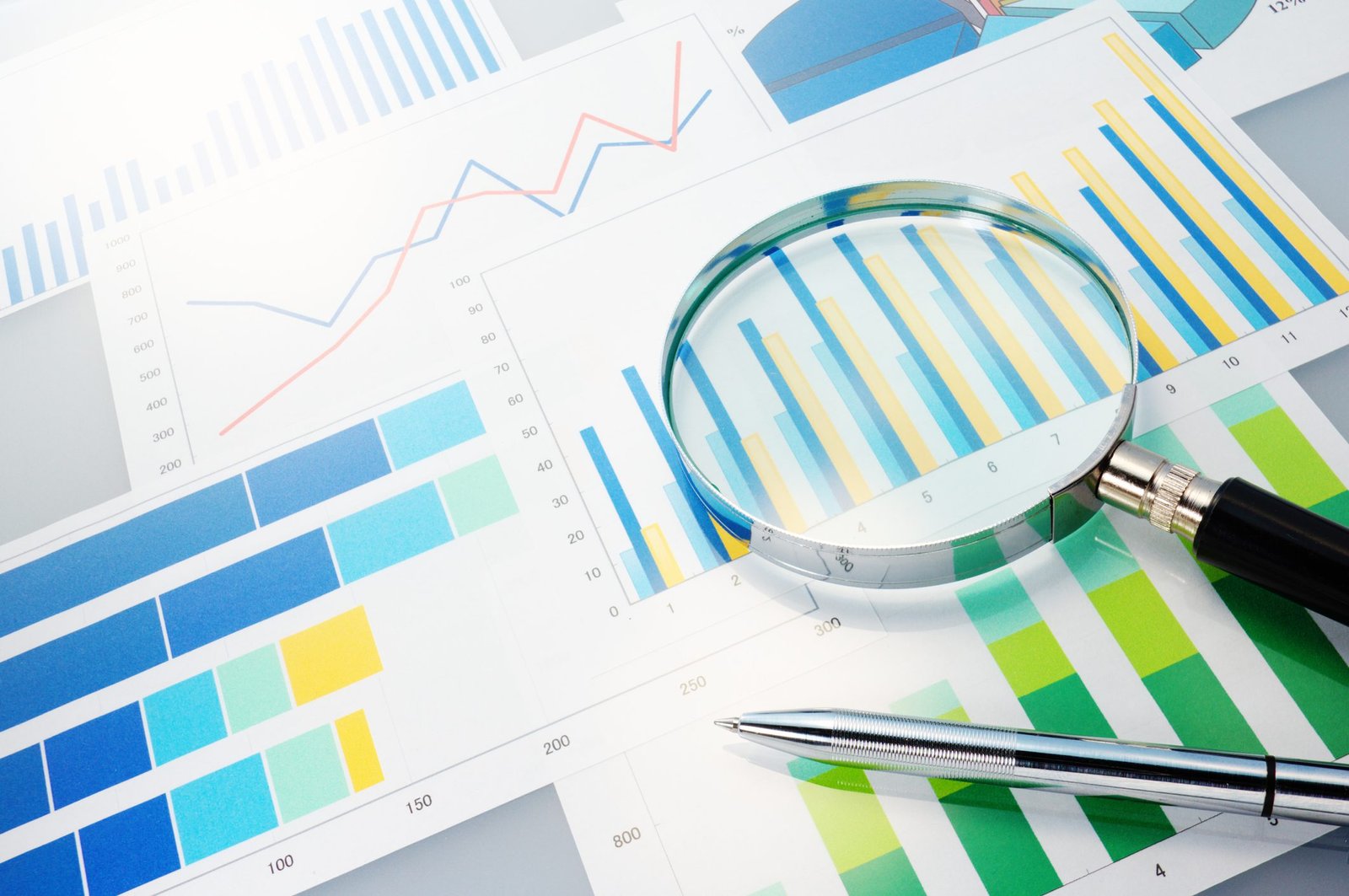 A magnifying glass and pen rest on various graphs and charts. The documents feature colorful bar graphs, line graphs, and pie charts, depicting data analysis. Sunlight illuminates the papers, casting soft shadows.